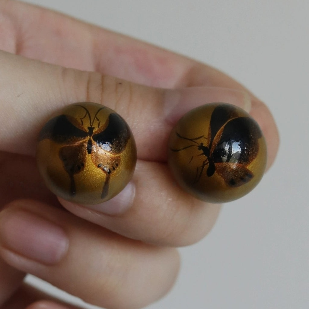 Two round, brown objects with black insect-like designs held between fingers against a neutral background