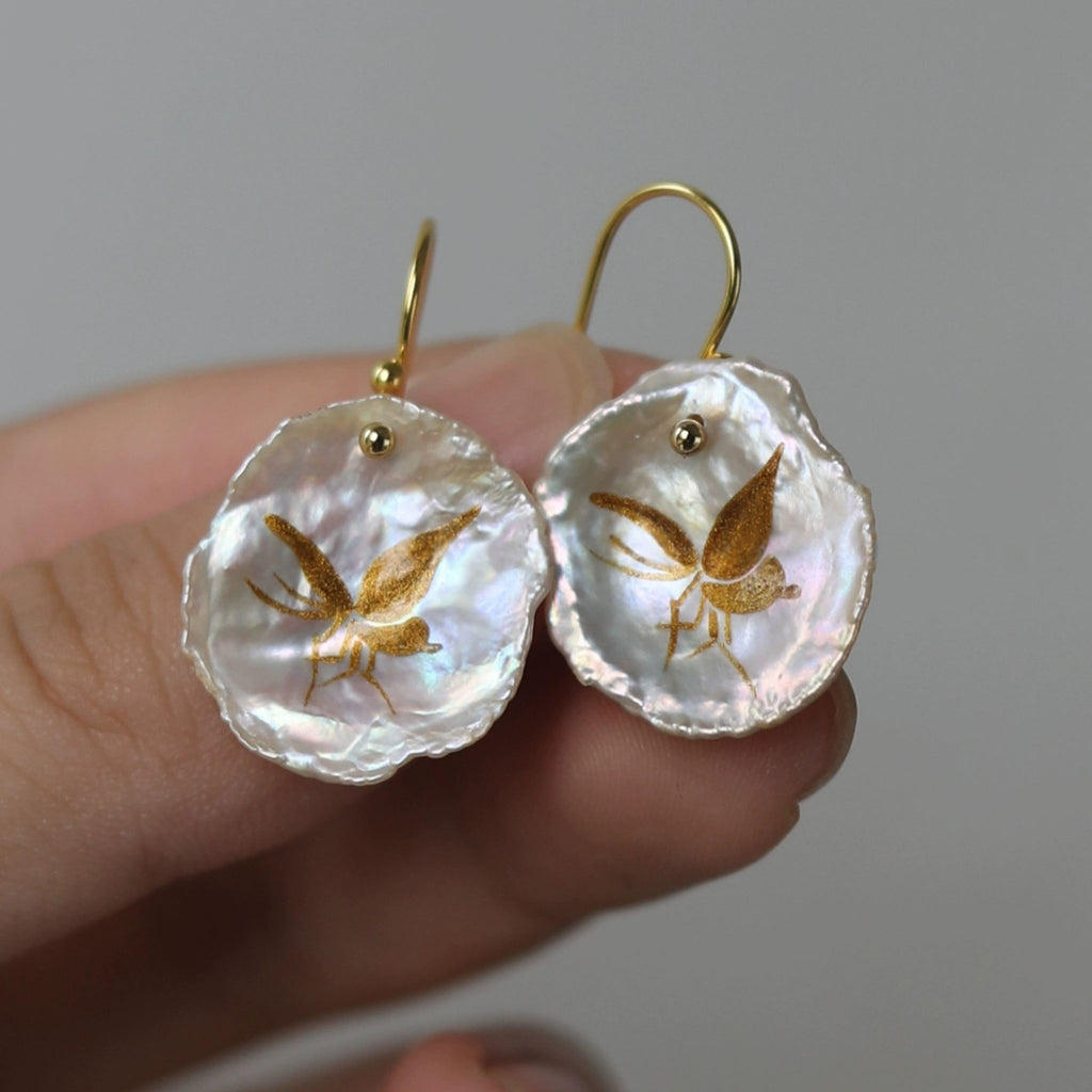 Pearl earrings with gold butterfly design held by a hand against a neutral background