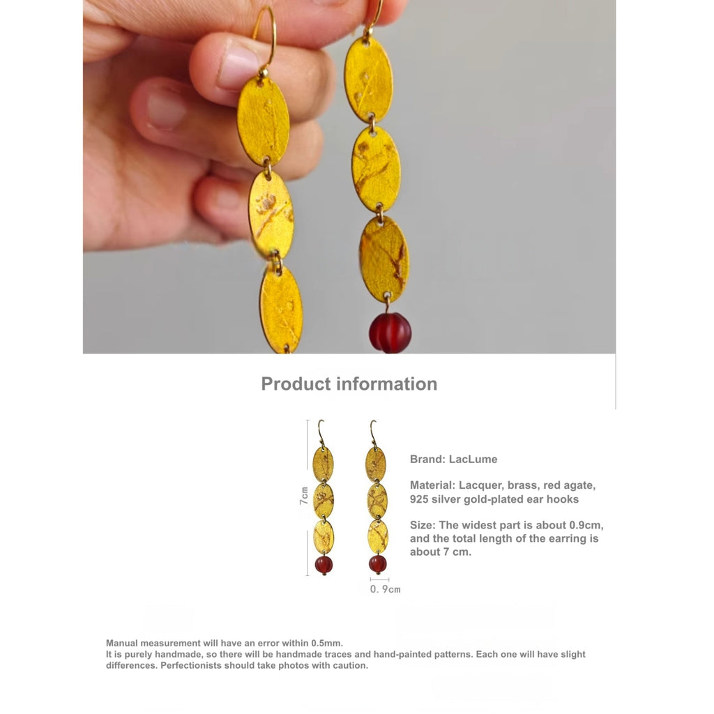 Yellow earrings with red accents held by a hand, displayed on a gray background with product information text.