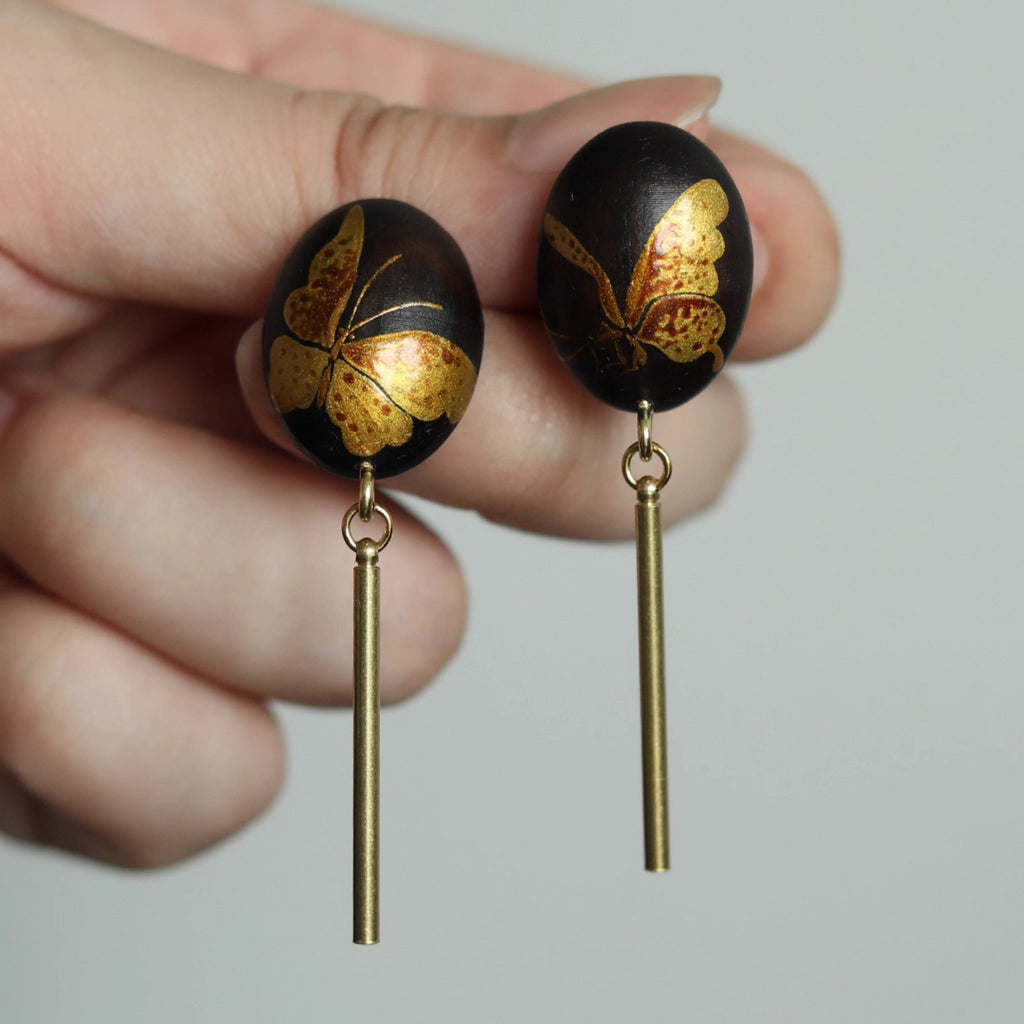 Black earrings with gold butterfly designs held between fingers against a neutral background LacLume Hand-Painted Lacquer Earrings Gold Butterfly Glamour Understated Elegance