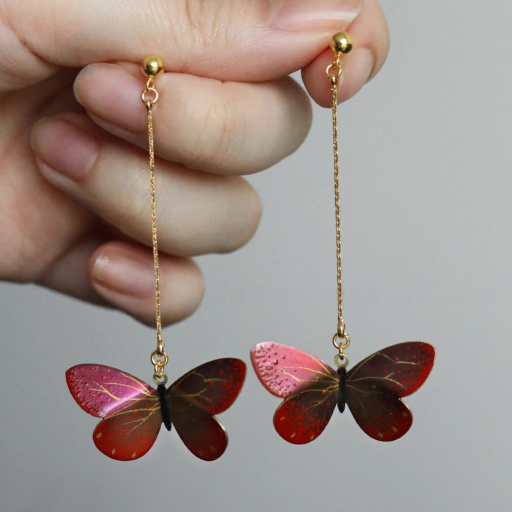 Butterfly-shaped earrings on gold chains held by a hand against a neutral background