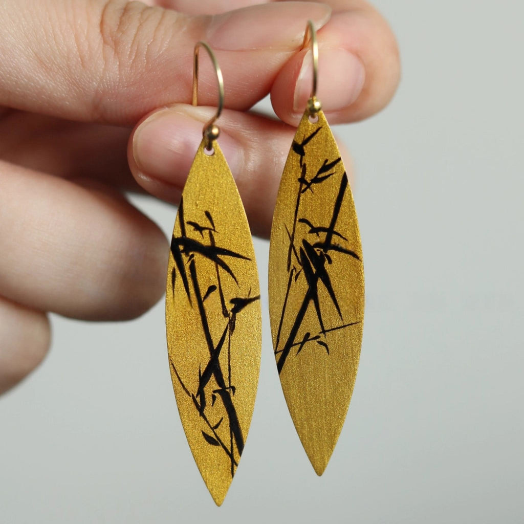 Gold earrings with black bamboo design held by a hand against a gray background