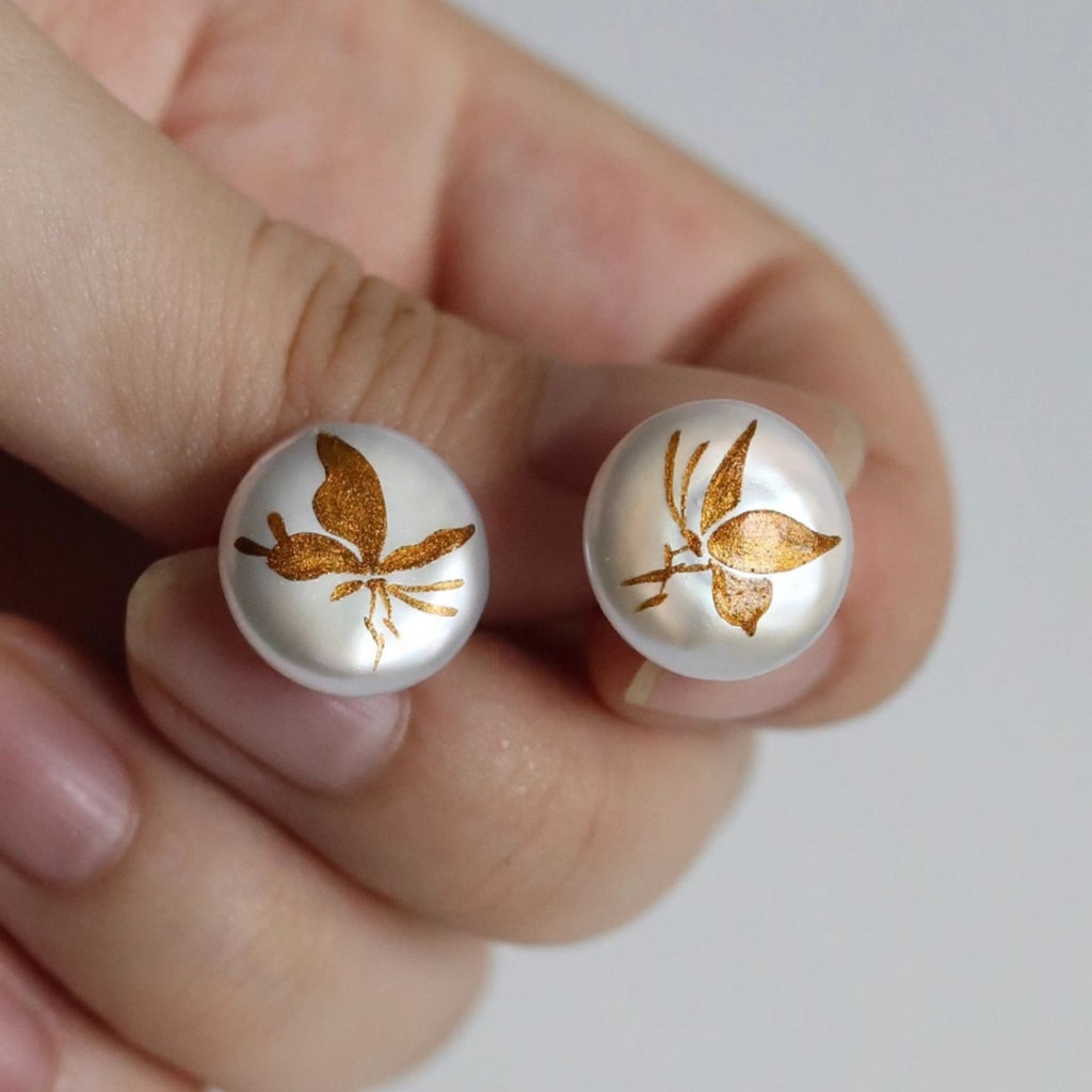 Two round earrings with butterfly design held between fingers against a neutral background LacLume Hand-Painted Lacquer Earrings Baroque Pearl Gilded Butterfly