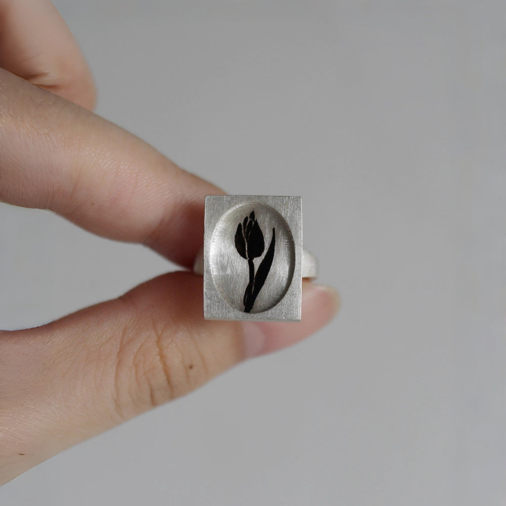 Silver ring with a black tulip design held by a hand against a neutral background