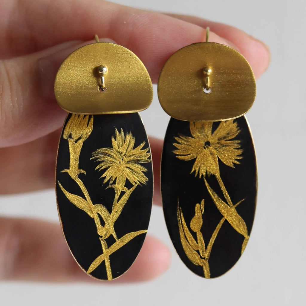 Gold and black floral earrings held between fingers against a neutral background