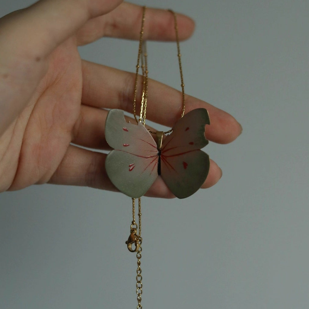 Butterfly-shaped pendant on a chain held by a hand against a neutral background