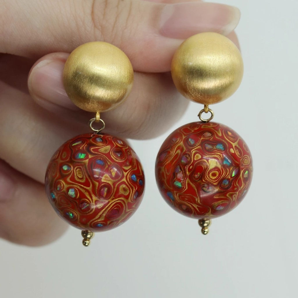 Gold and red patterned earrings held between fingers against a white background