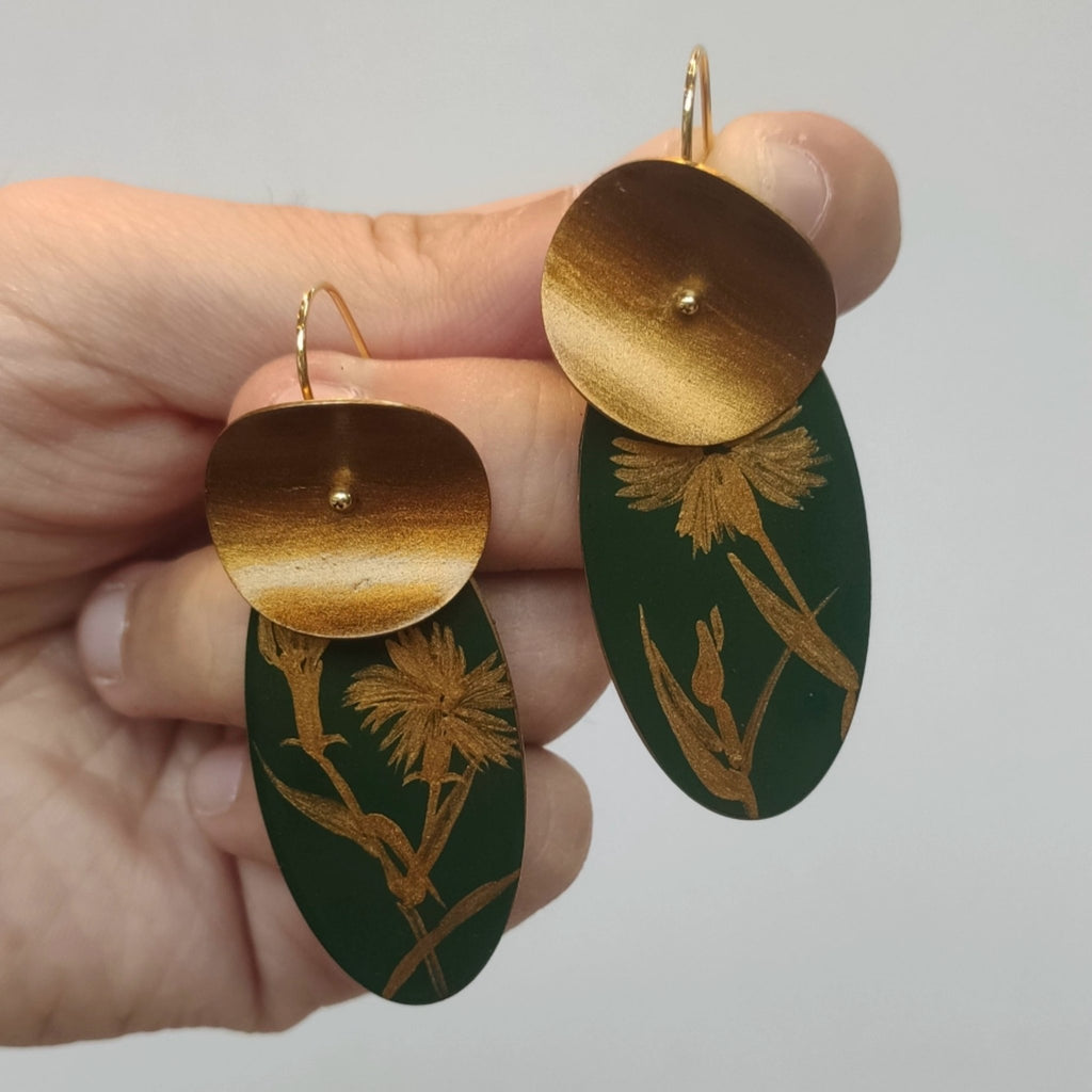 Hand holding green and gold earrings with floral design against a gray background