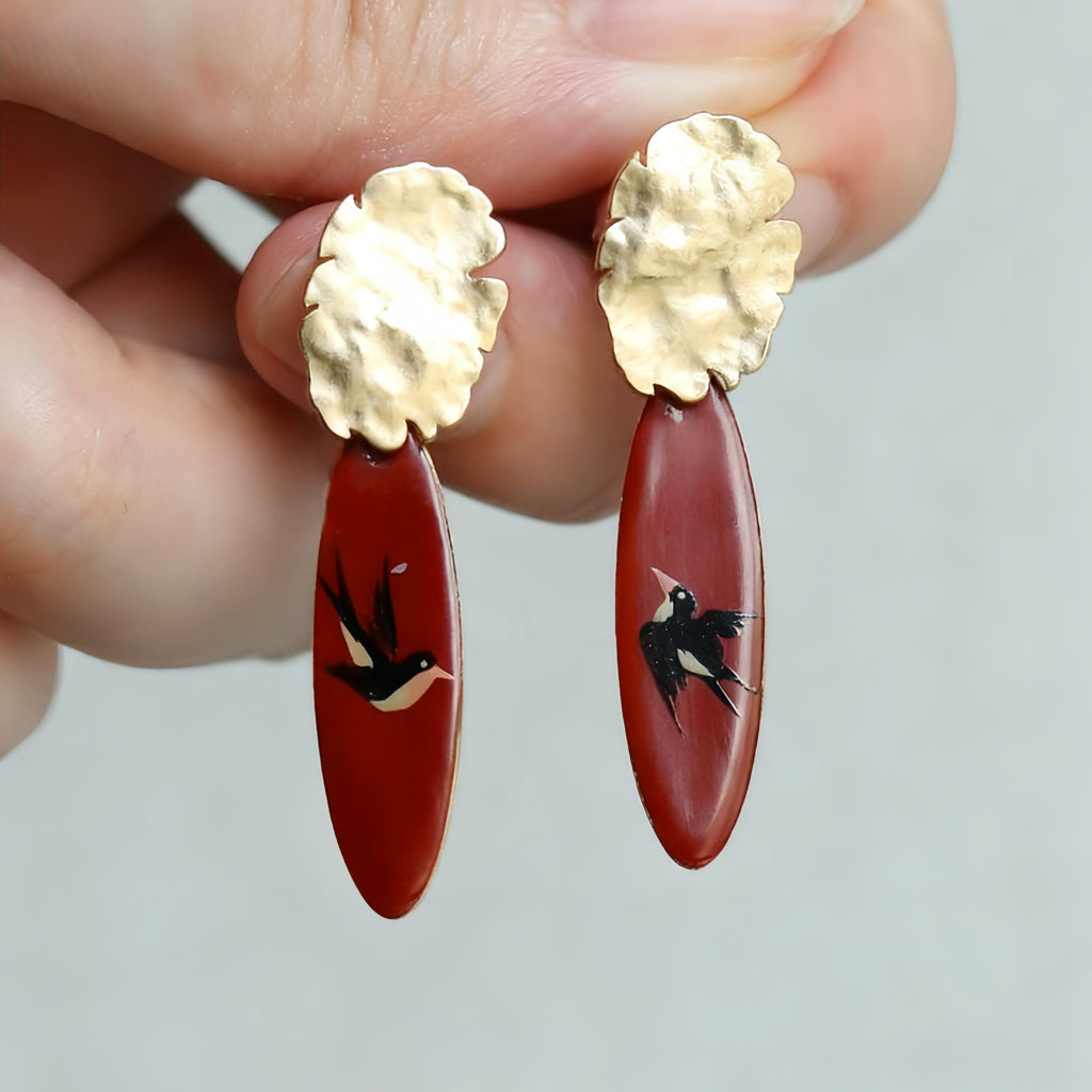 Gold and red earrings with bird designs held by a hand against a neutral background