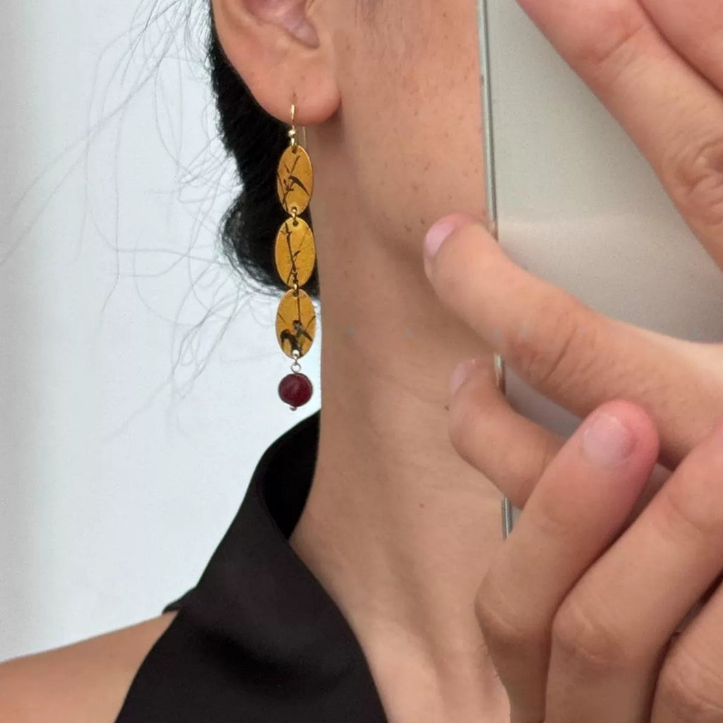 Close-up of a person wearing gold earrings with maroon beads, holding a phone.