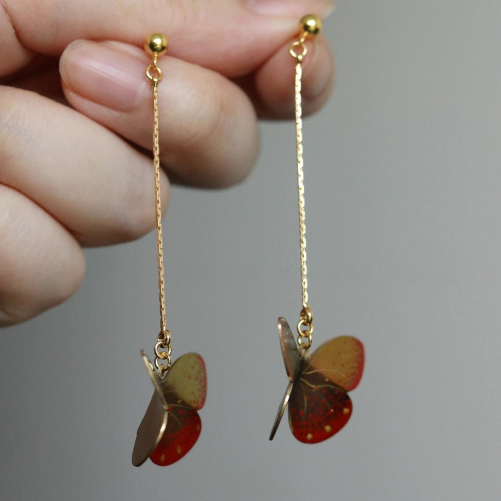 Butterfly-shaped earrings with gold chains held by a hand against a neutral background