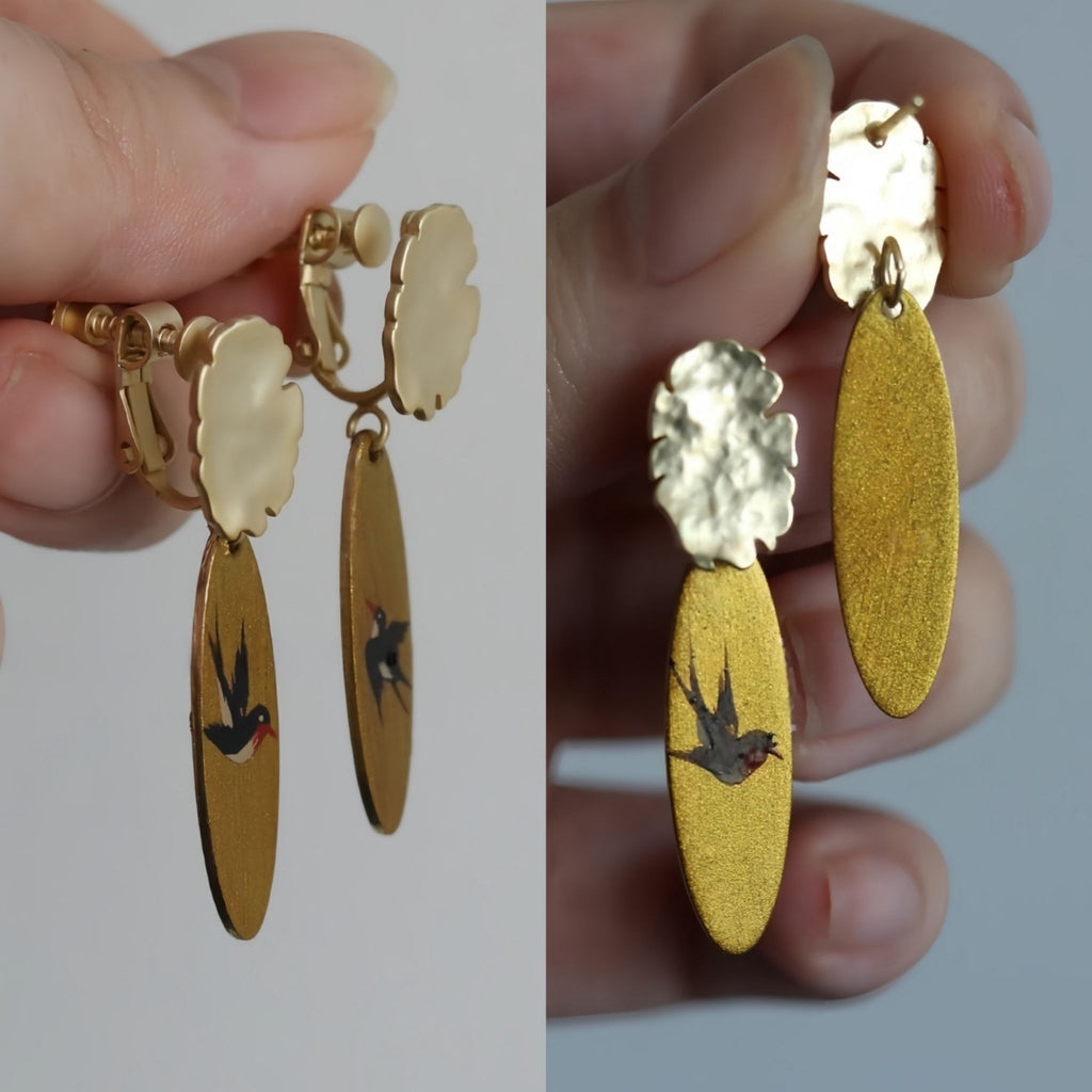Gold earrings with yellow oval pendants and bird designs held by a hand.