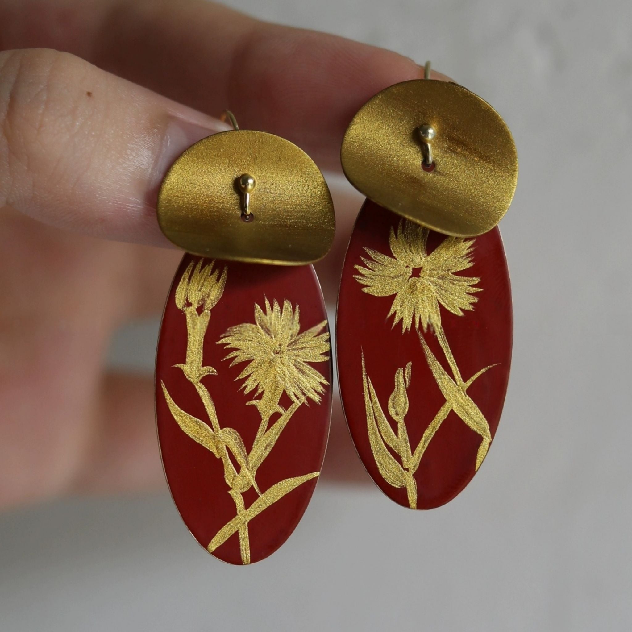 Gold floral earrings on red oval stones held by a hand against a neutral background