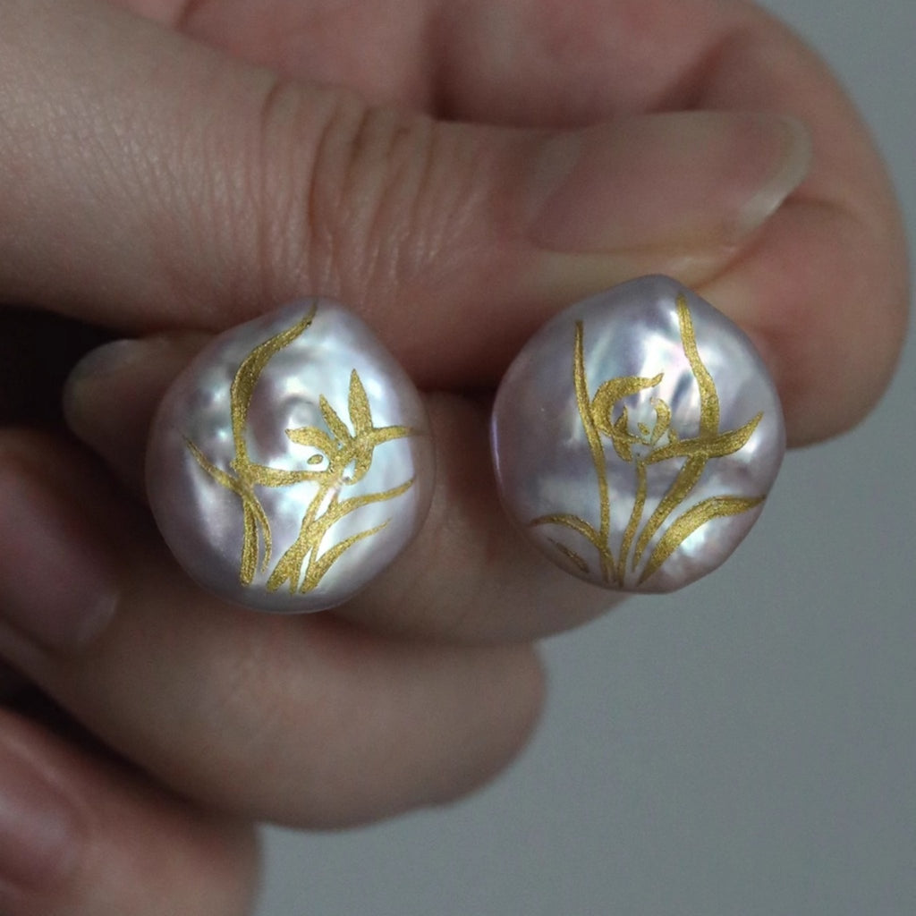 Pearl earrings with gold floral patterns held between fingers against a neutral background