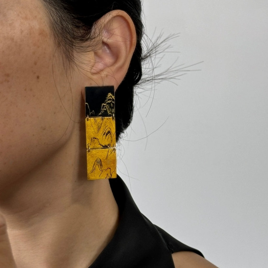 Close-up of a person wearing a black and gold square earring against a neutral background LacLume Retro Style Lacquer Earrings HandPaintedd Mountain Peaks