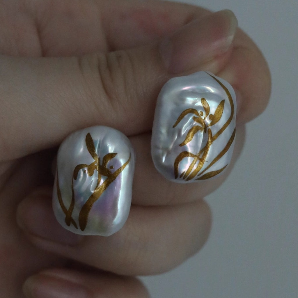 Pearl earrings with gold floral designs held between fingers against a neutral background