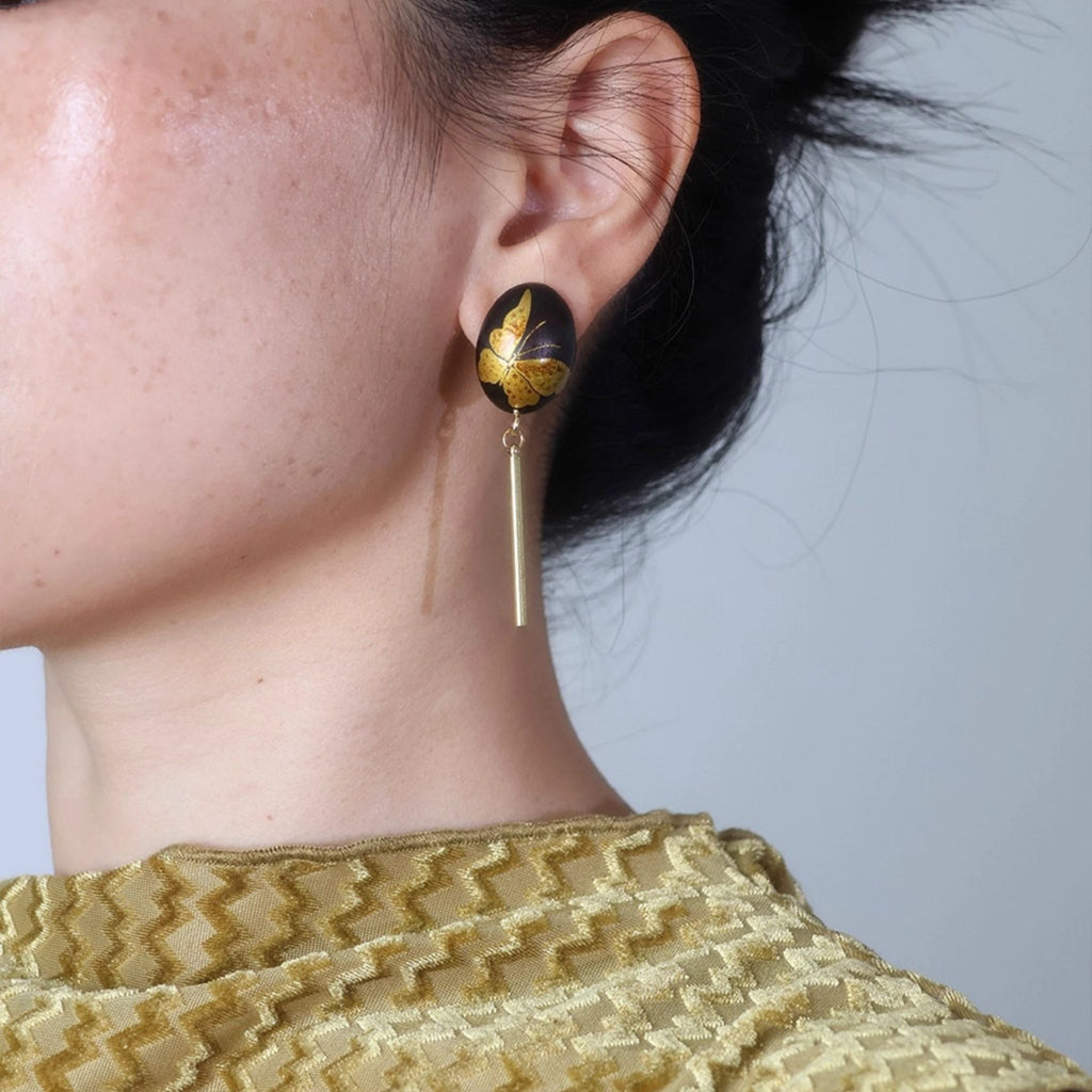 Close-up of an ear wearing a decorative earring with a gold and black design. LacLume Hand-Painted Lacquer Earrings Gold Butterfly Glamour Understated Elegance