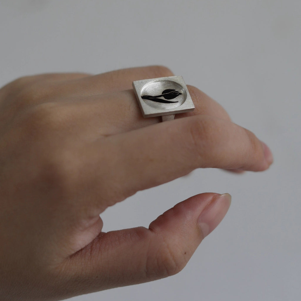 Silver ring with a black design on a person's finger against a plain background
