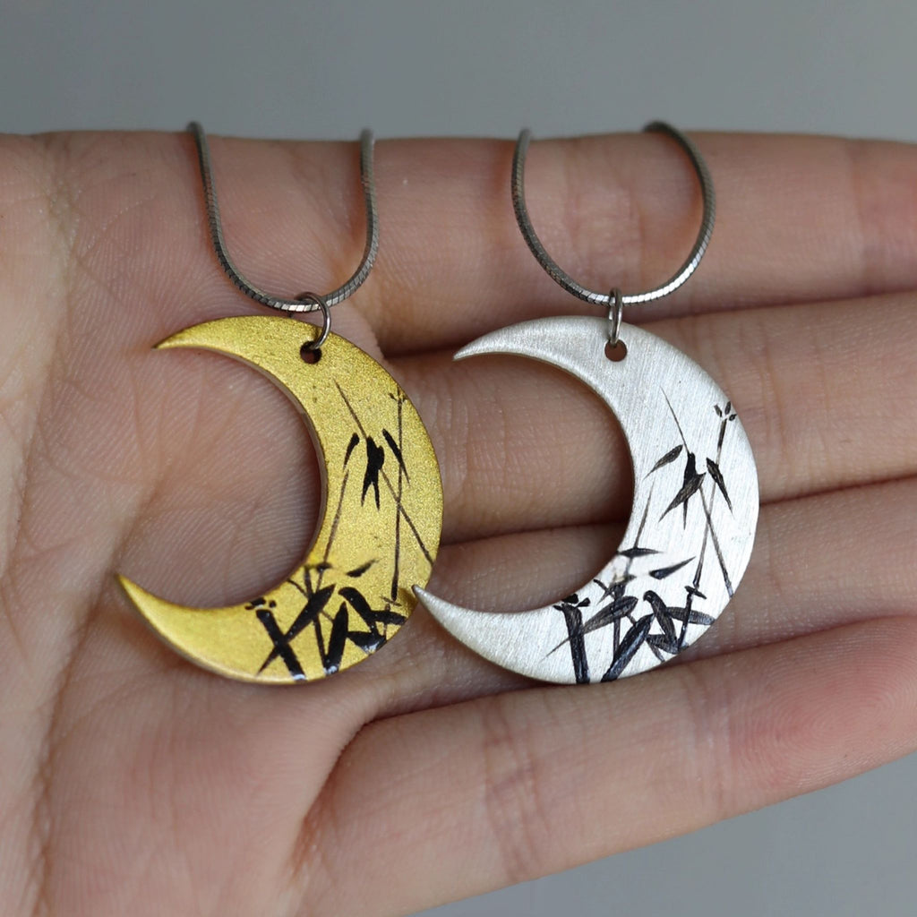 Two moon-shaped pendants, one gold and one silver, held in a hand against a neutral background.