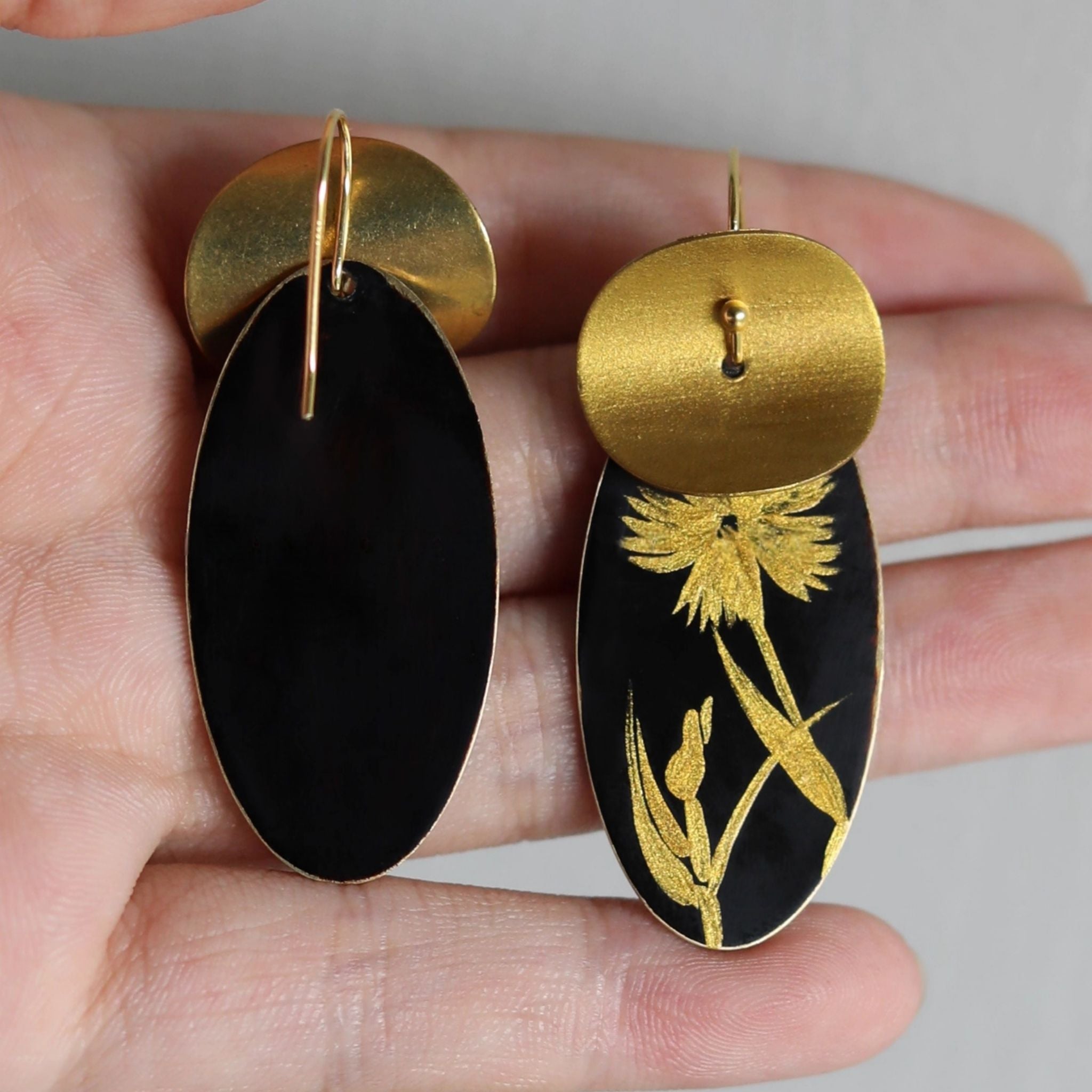 Gold and black earrings with floral design held in a hand against a neutral background