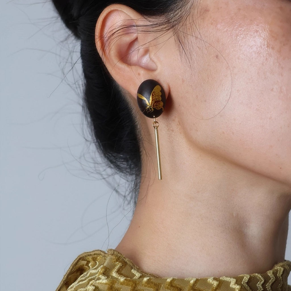 Close-up of a person wearing a black and gold earring with a gold tag against a neutral background LacLume Hand-Painted Lacquer Earrings Gold Butterfly Glamour Understated Elegance