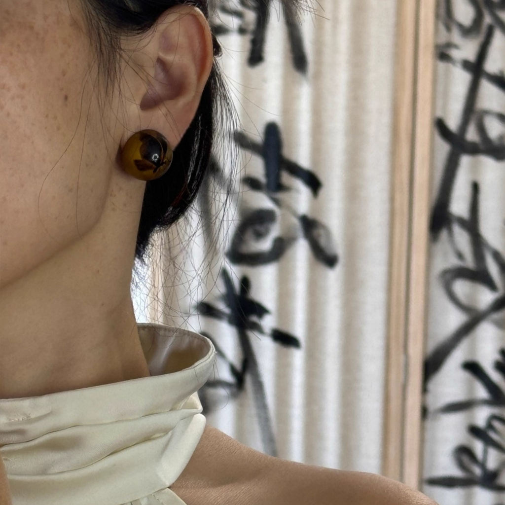 Close-up of a person wearing a brown circular earring with a blurred  LacLume HandPainted Retro Autumn Butterfly Earrings Luxe Artisanal Lacquer Jewelrybackground